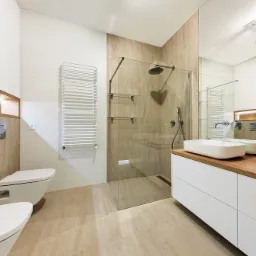 Nowoczesna łazienka z beżowymi płytkami imitującymi kamień, kabiną prysznicową typu walk-in z czarną armaturą, podwieszaną miską WC i bidetem, oraz białą szafką z drewnianym blatem pod umywalki.