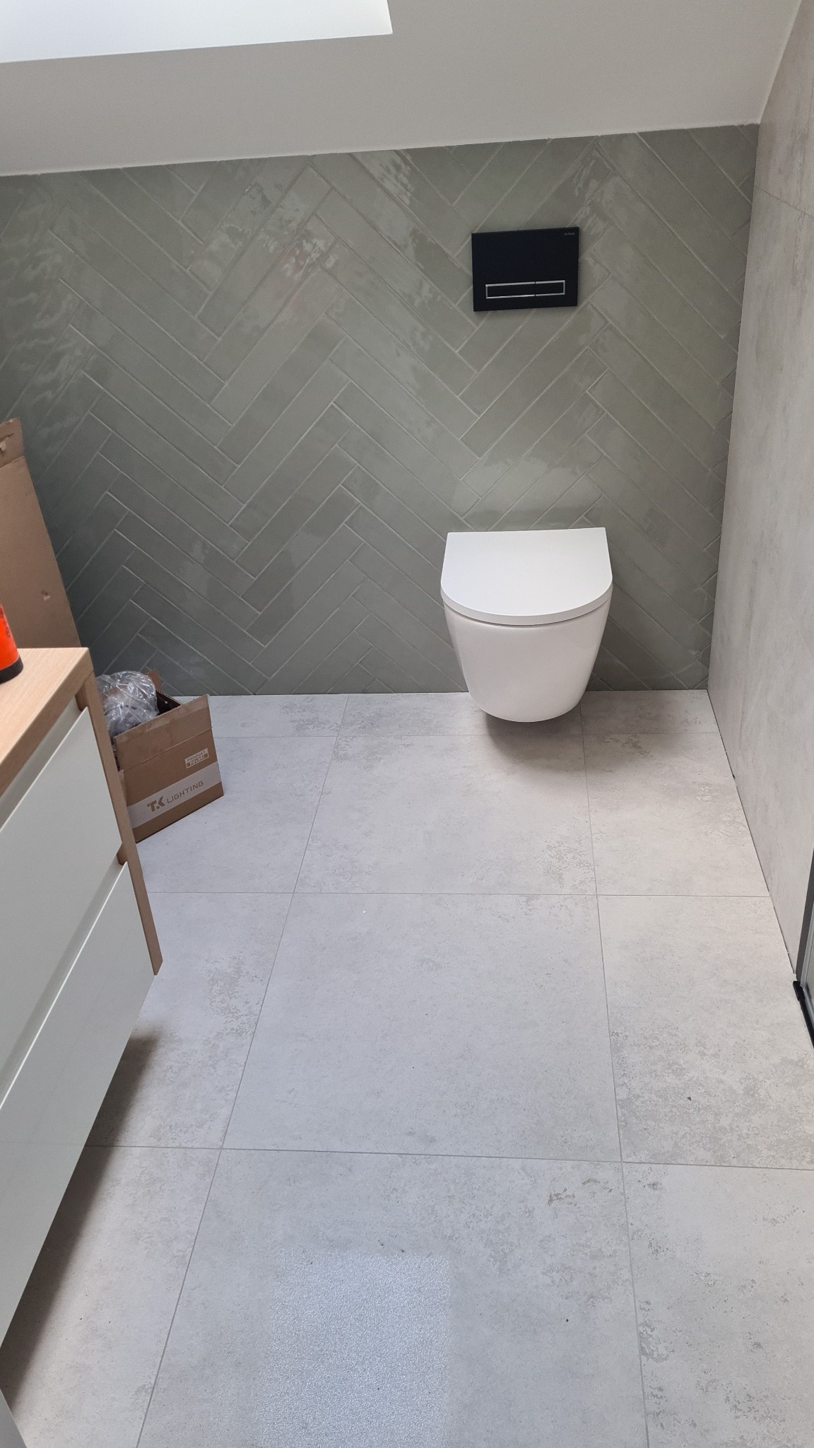 Nowoczesna łazienka z wiszącą toaletą, ściana wyłożona zielonkawą glazurą w jodełkę, jasne płytki podłogowe, częściowo widoczna szafka z umywalką i karton po akcesoriach.