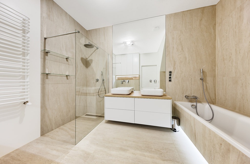 Jasna łazienka z beżowymi płytkami imitującymi marmur, kabiną prysznicową typu walk-in, podwójną umywalką nablatową na białej szafce z drewnianym blatem oraz grzejnikiem drabinkowym.
