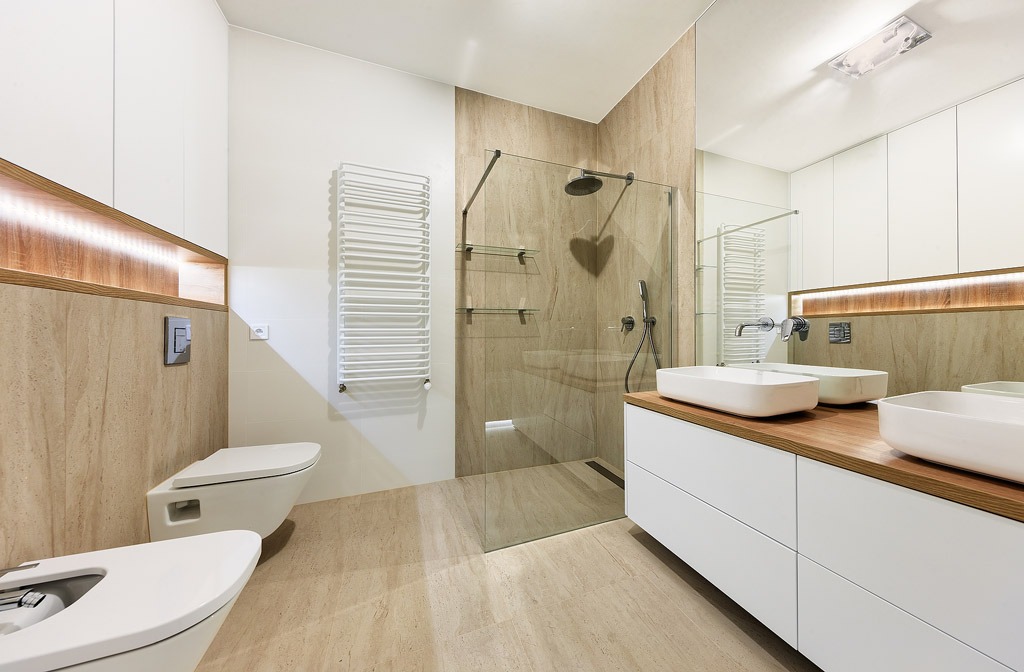 Nowoczesna łazienka z beżowymi płytkami imitującymi kamień, kabiną prysznicową typu walk-in z czarną armaturą, podwieszaną miską WC i bidetem, oraz białą szafką z drewnianym blatem pod umywalki.