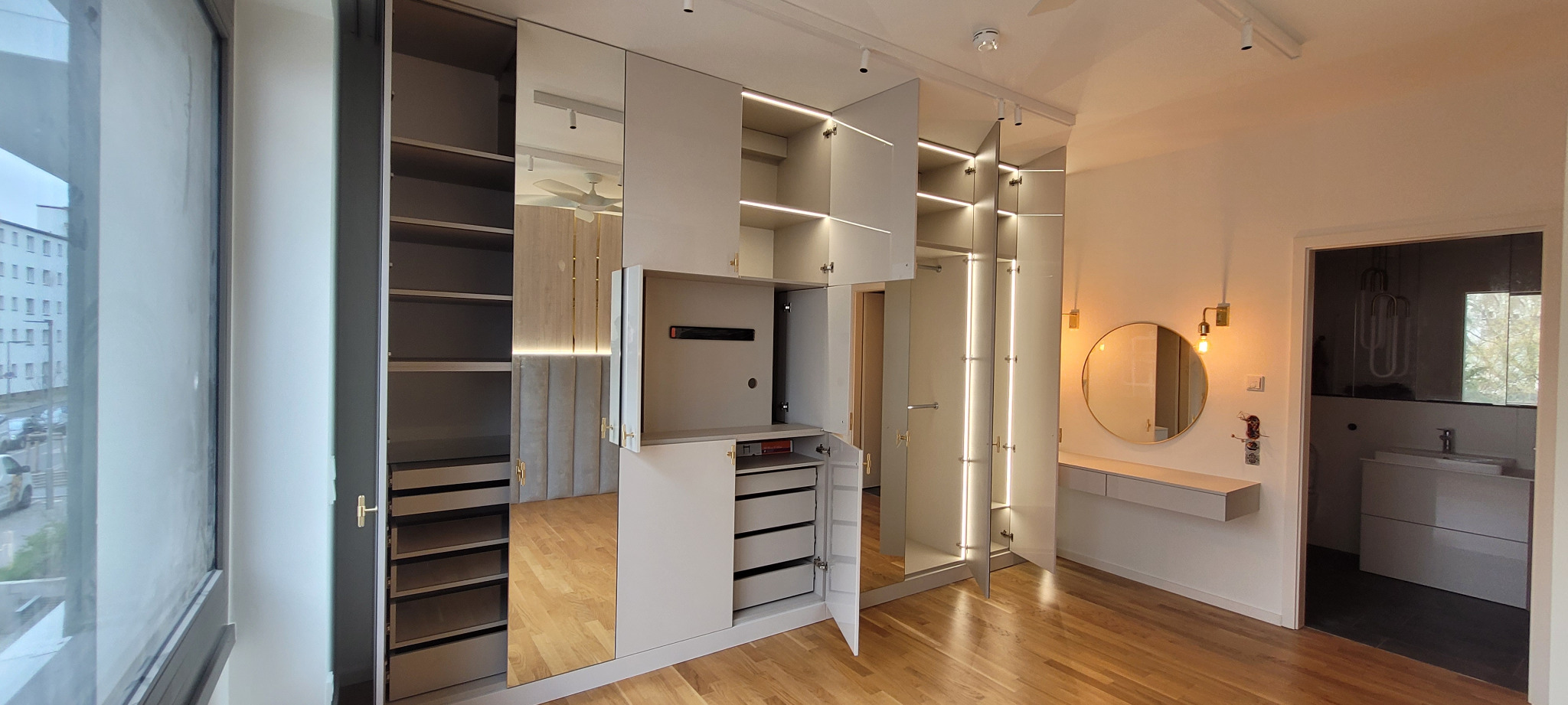 Jasna szafa wnękowa z lustrzanymi drzwiami i oświetleniem LED w nowoczesnej sypialni z drewnianą podłogą, widoczne otwarte półki i szuflady.