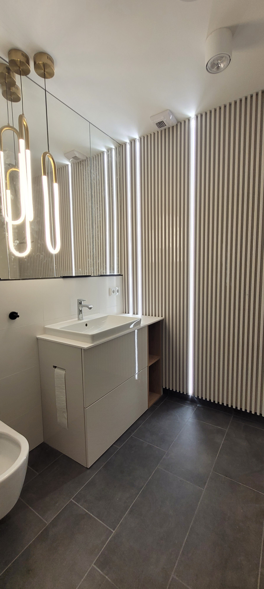 Nowoczesna łazienka z białym blatem umywalkowym, szarą podłogą z płytek i ścianą z pionowych paneli z podświetleniem LED.