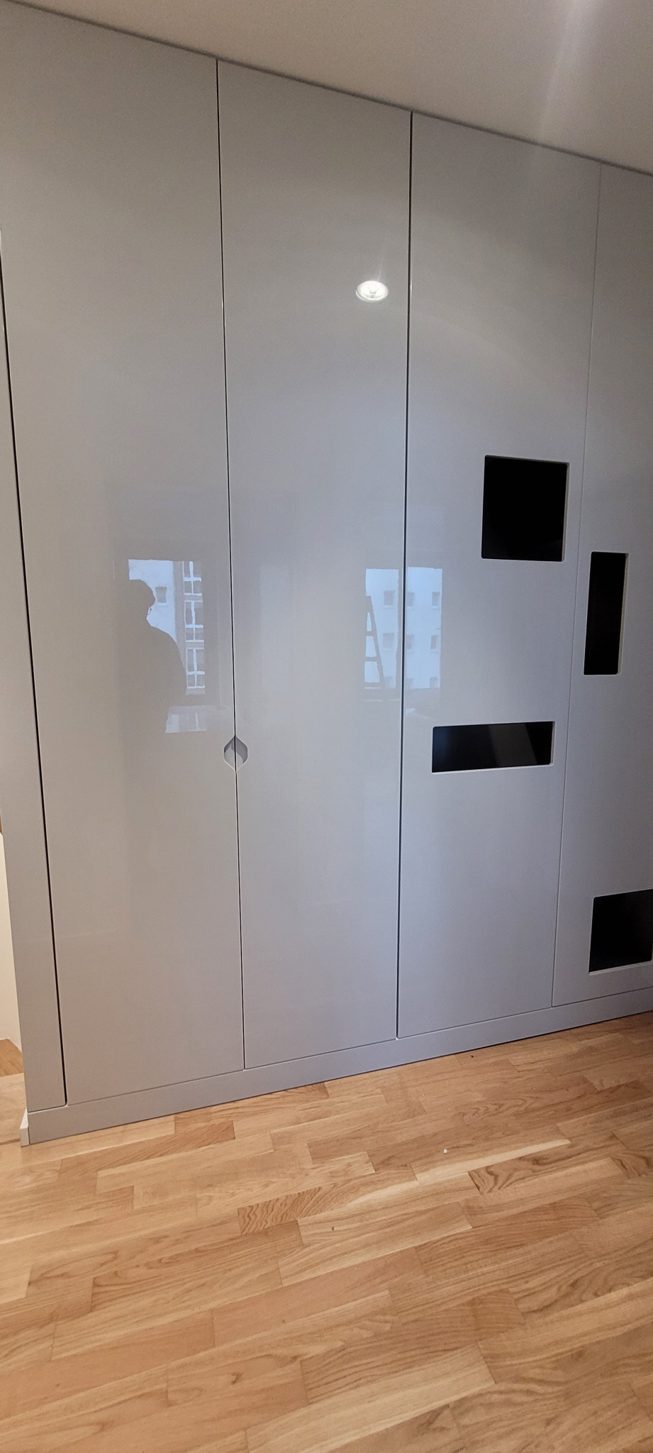 Szara zabudowa meblowa z połyskującymi frontami i małymi, prostokątnymi otworami wentylacyjnymi, wbudowana we wnękę na podłodze wyłożonej parkietem.