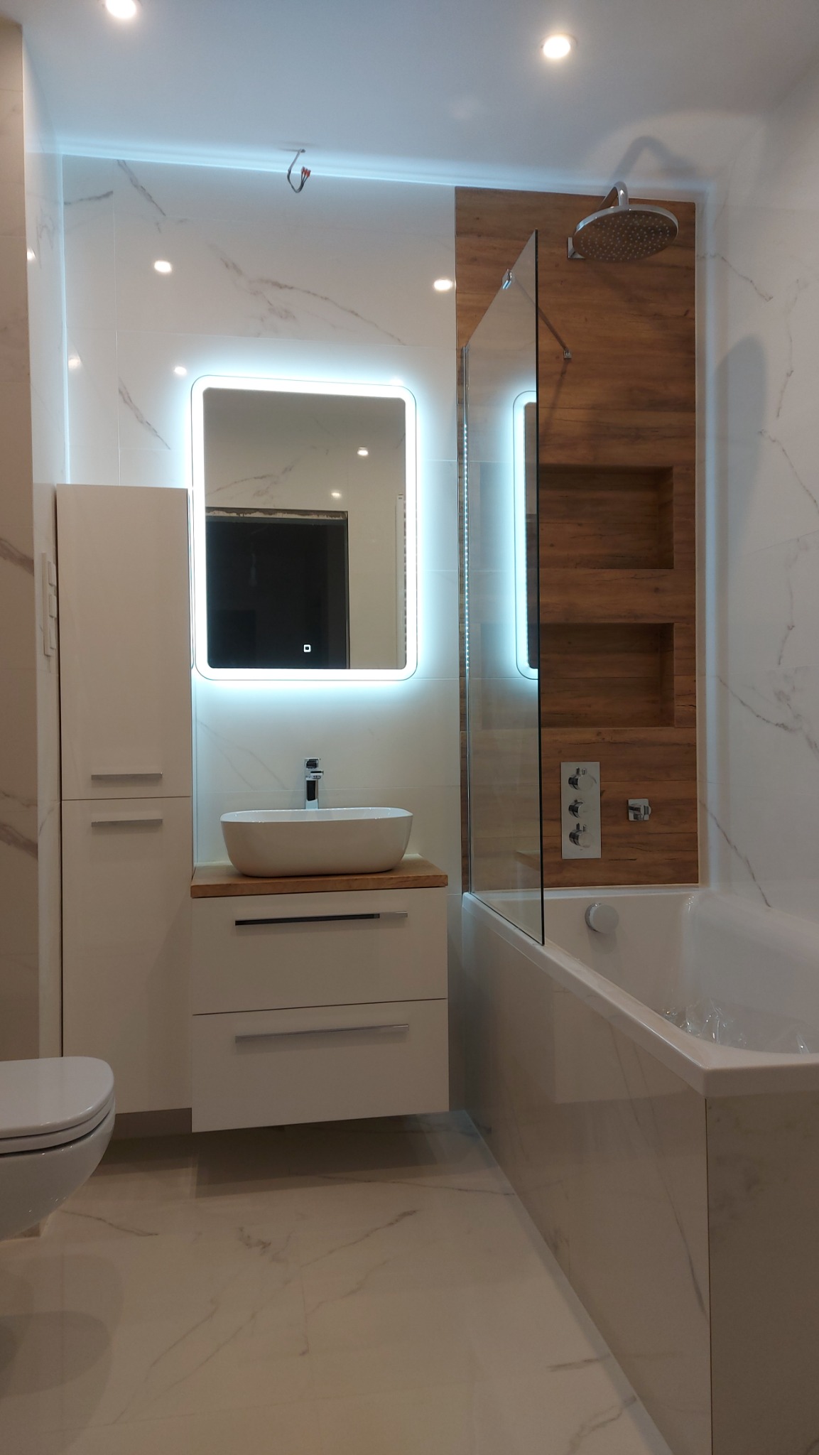 Nowoczesna łazienka z podświetlanym lustrem, umywalką nablatową i kabiną prysznicową za szklaną ścianką, wykończona płytkami imitującymi marmur i drewno.
