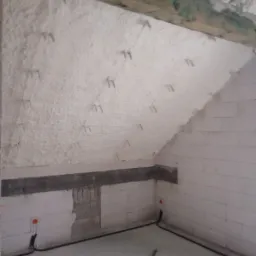 Poddasze ocieplone białą pianką PUR z widocznymi mocowaniami, surowe ściany z białego bloczka, czarne przewody elektryczne wzdłuż podłogi.