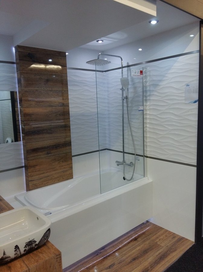 Nowoczesna łazienka z wanną obudowaną białą płytą, szklanym parawanem nawannowym i deszczownicą, ściany wykończone białymi płytkami 3D oraz panelami imitującymi drewno, podłoga wyłożona panelami...