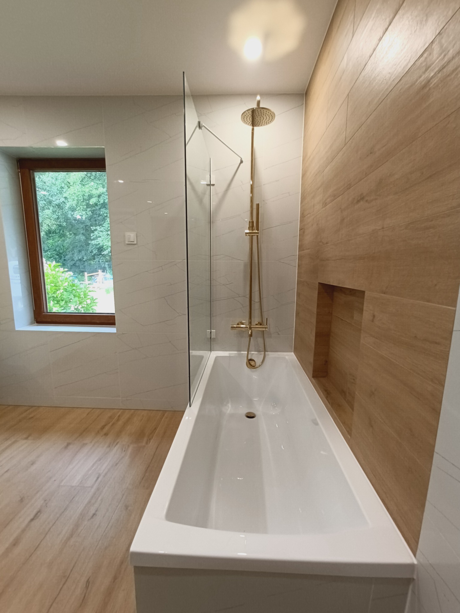 Nowoczesna łazienka z wanną, złotą armaturą prysznicową i szklanym parawanem, wykończona płytkami imitującymi drewno i marmur, z widokiem na zieleń za oknem.