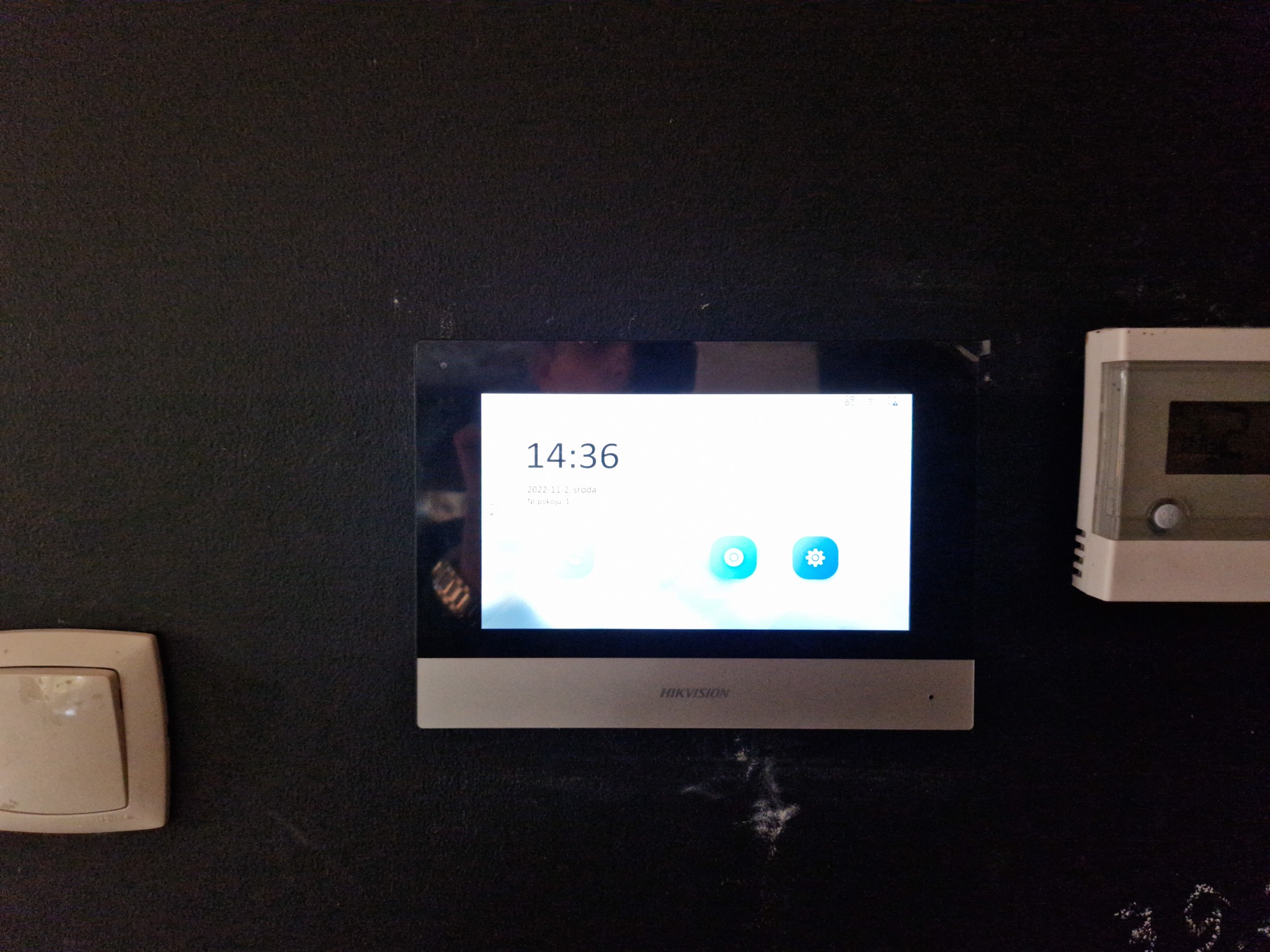 Panel sterowania wideodomofonu Hikvision zamontowany na ciemnej ścianie, obok widoczny termostat i włącznik światła