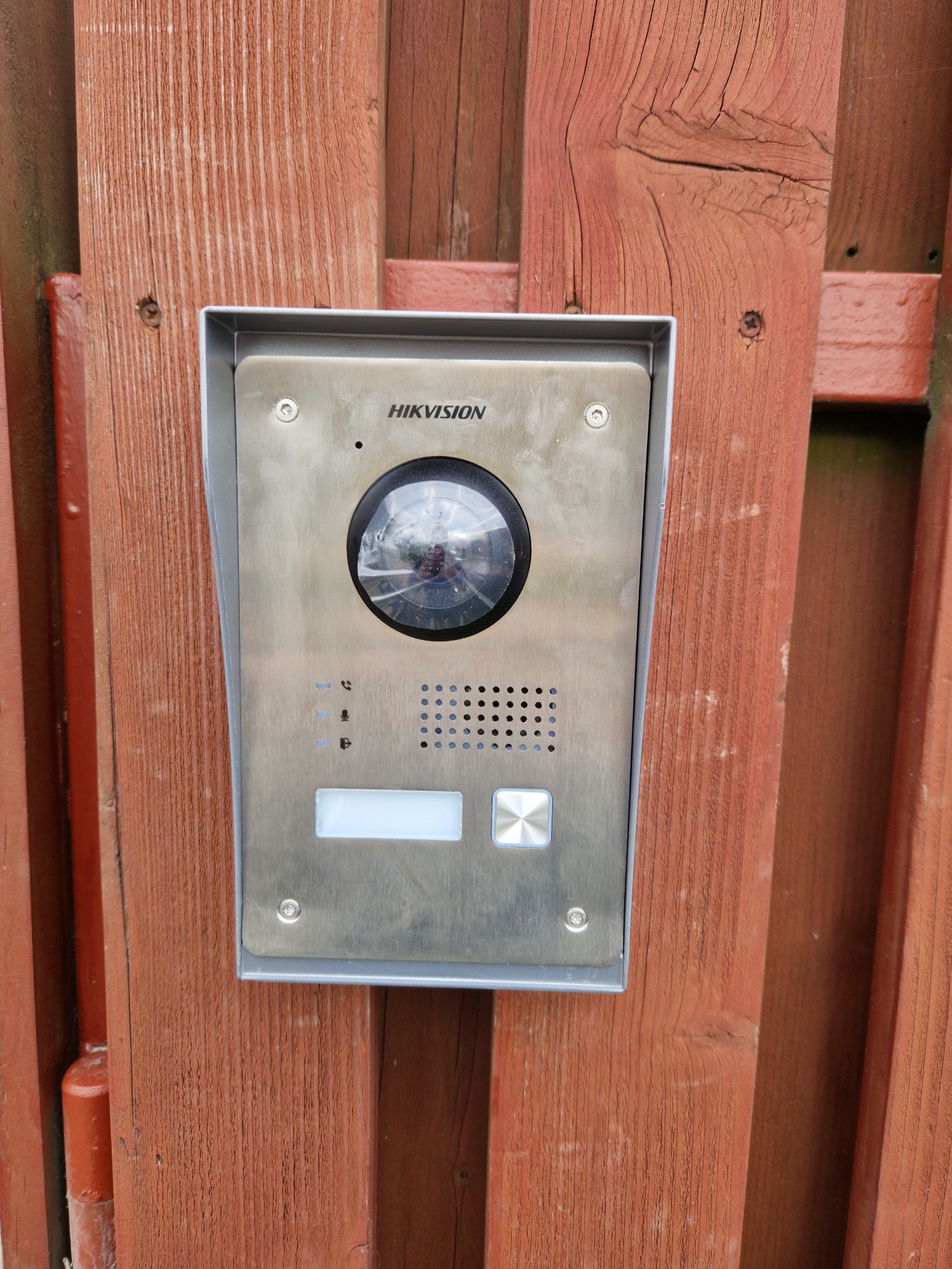 Zewnętrzny panel wideodomofonu Hikvision zamontowany na drewnianym ogrodzeniu z widocznymi śrubami mocującymi.