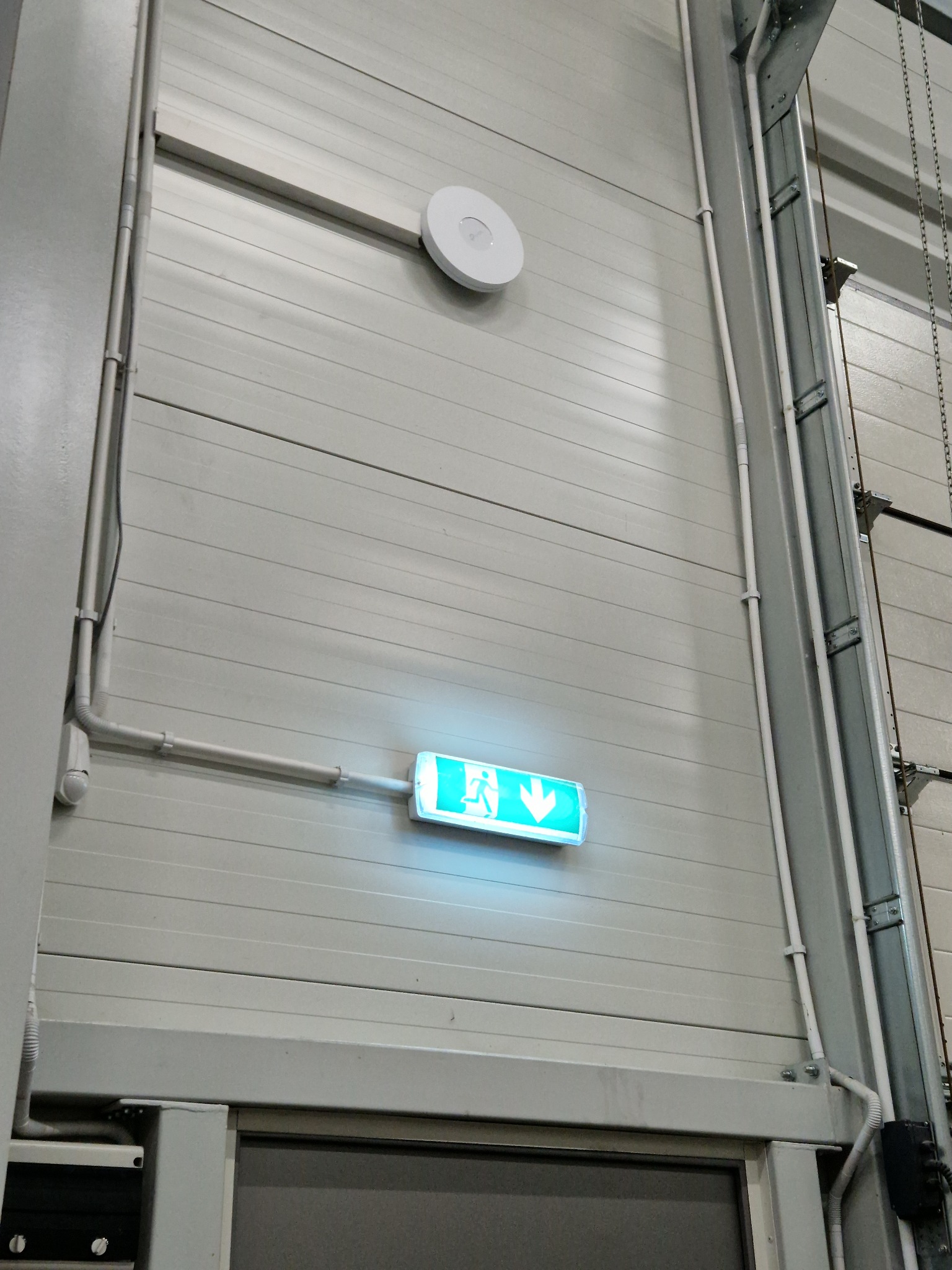 Świecący, zielony znak ewakuacyjny z piktogramem osoby biegnącej w dół, zamontowany na panelowej ścianie w hali, z widocznymi przewodami i czujnikiem dymu.