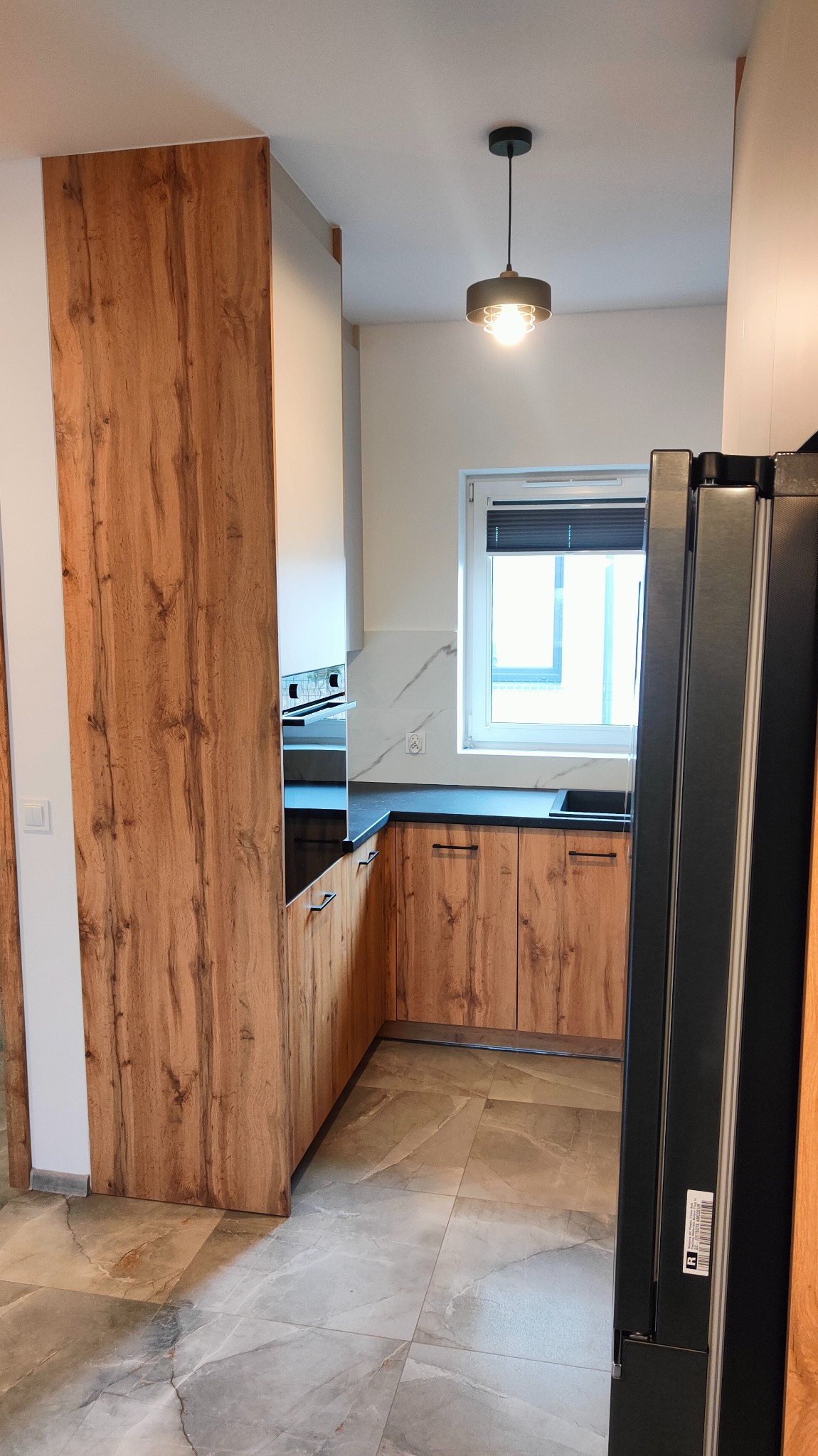 Widok na nowoczesną kuchnię z drewnianymi frontami szafek, czarnym blatem i marmurowymi płytkami na ścianie, oświetloną wiszącą lampą z ozdobnym żarnikiem.