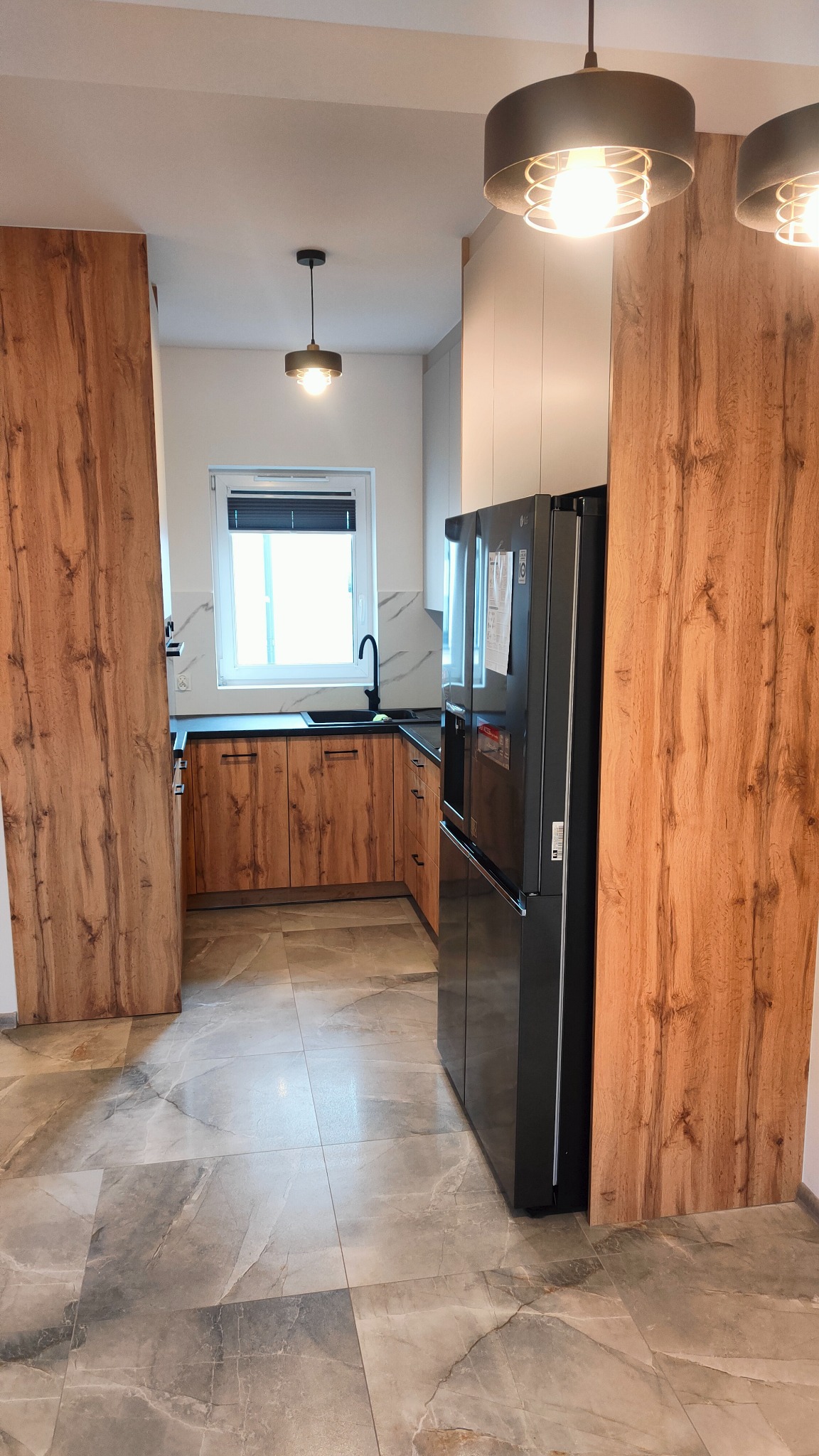 Nowoczesna kuchnia z drewnianymi frontami szafek, czarnym blatem i zlewozmywakiem, marmurową podłogą oraz dużą, czarną lodówką z wyświetlaczem.