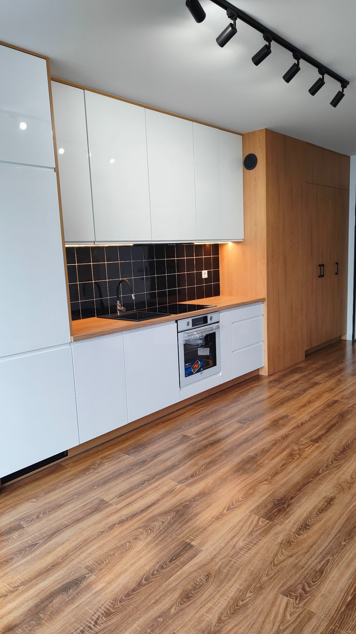 Nowoczesna kuchnia z białymi, lakierowanymi szafkami, drewnianym blatem, czarnymi płytkami nad blatem i drewnianą zabudową ściany z szafami, oświetlona reflektorami sufitowymi.