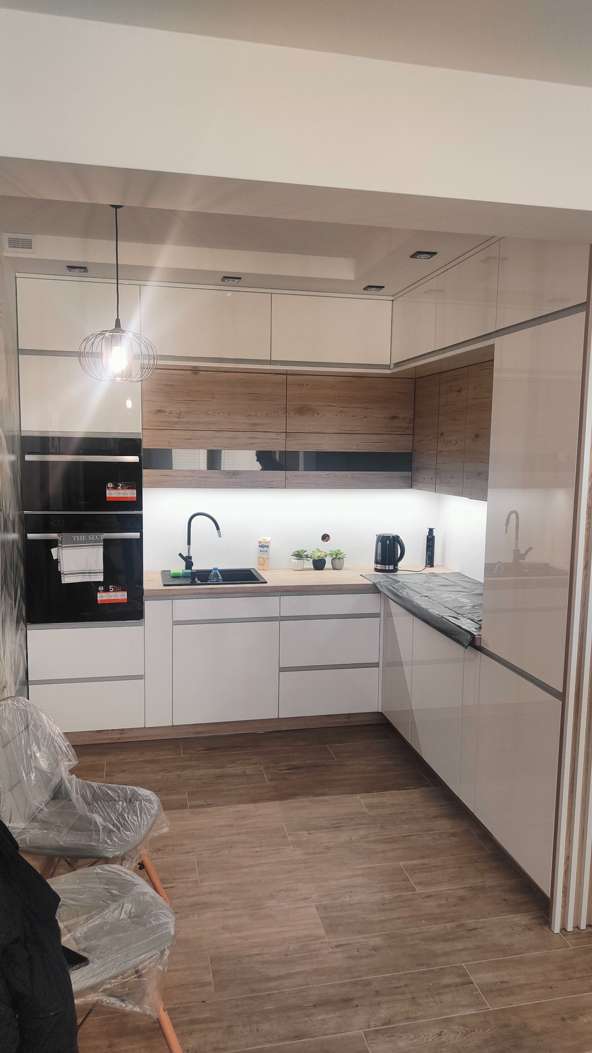 Nowoczesna kuchnia z białymi frontami i drewnianymi akcentami, czarnym zlewem i piekarnikiem, podświetleniem LED pod szafkami wiszącymi, wiszącą lampą drucianą i podłogą imitującą drewno.