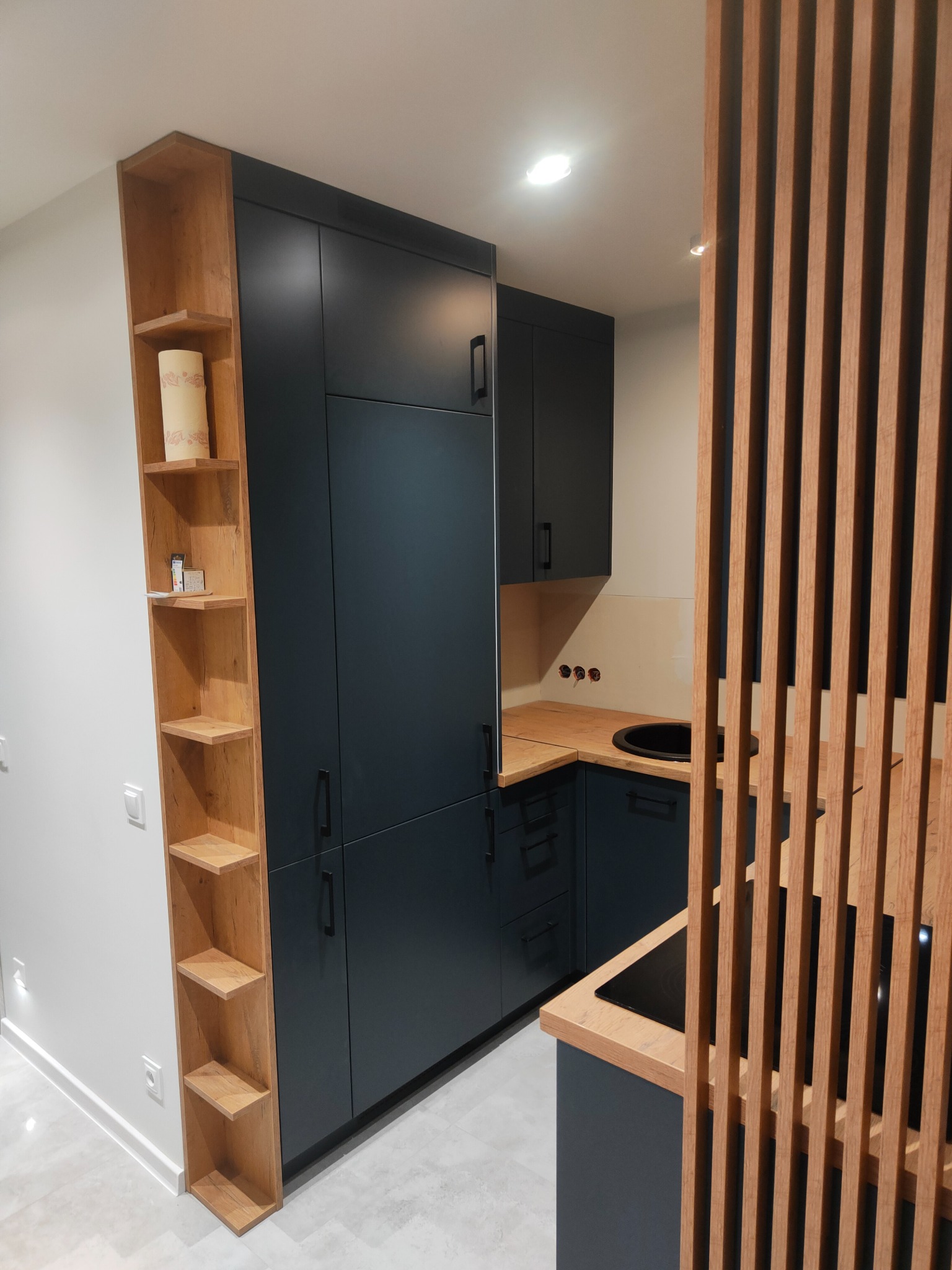 Nowoczesna, kompaktowa kuchnia w odcieniach granatu i drewna, z pionową drewnianą przesłoną oddzielającą przestrzeń. Wysoka szafka z półkami w kolorze drewna stoi obok zabudowy kuchennej. Blat...