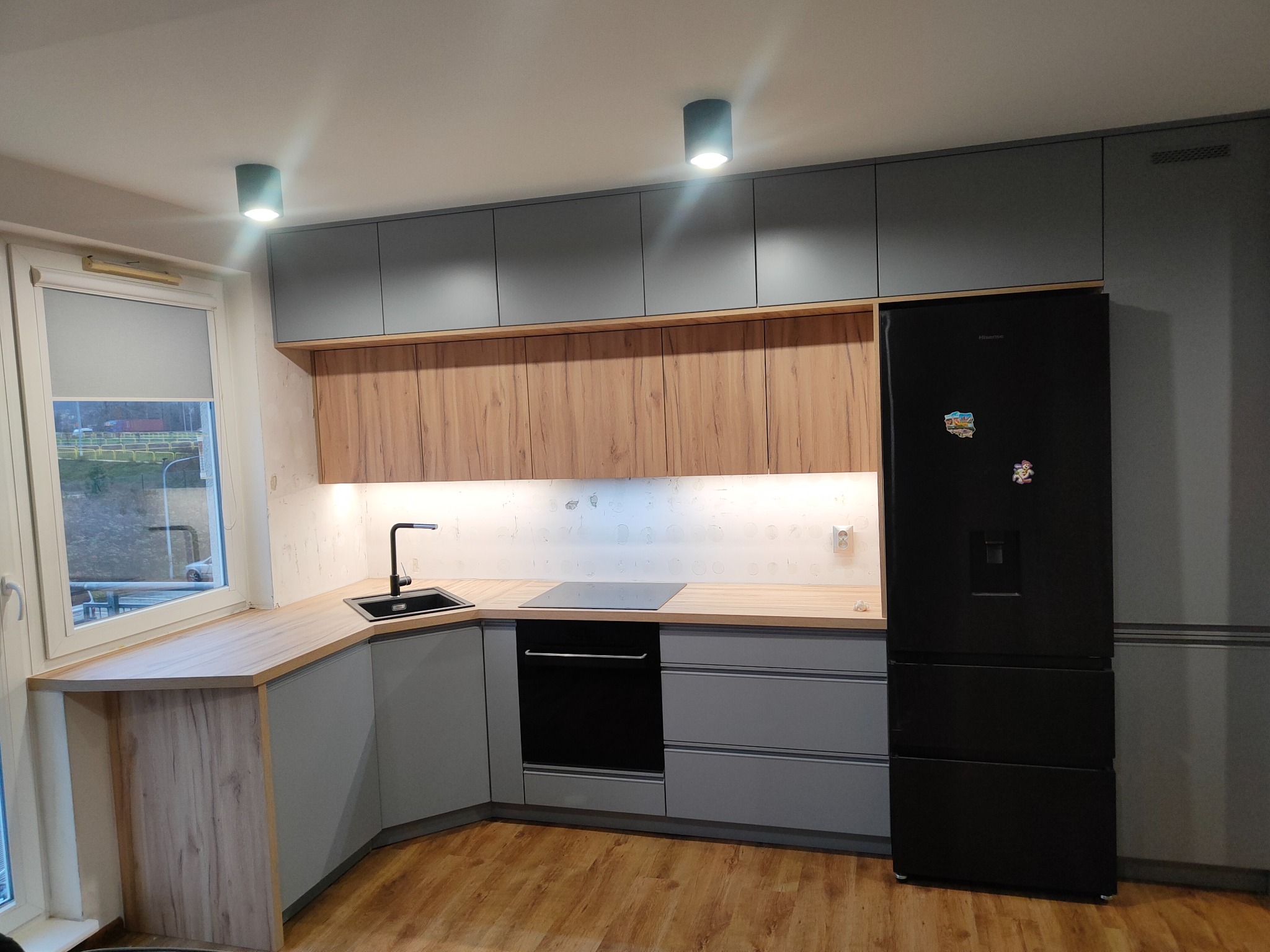 Nowoczesna kuchnia w odcieniach szarości i drewna, z czarnym zlewem, płytą indukcyjną i lodówką, oświetlona punktowymi lampami sufitowymi.