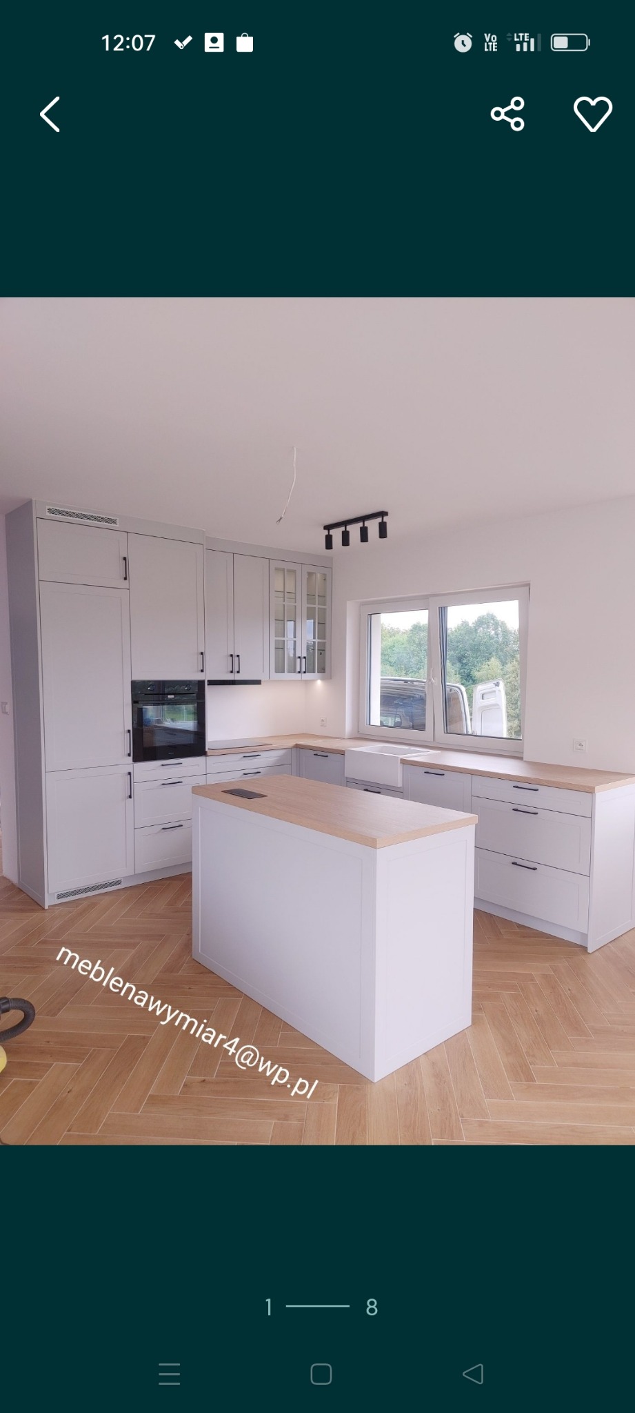 Jasna kuchnia na wymiar z wyspą i drewnianym blatem, podłoga ułożona w jodełkę, widoczne okno z zielenią na zewnątrz.