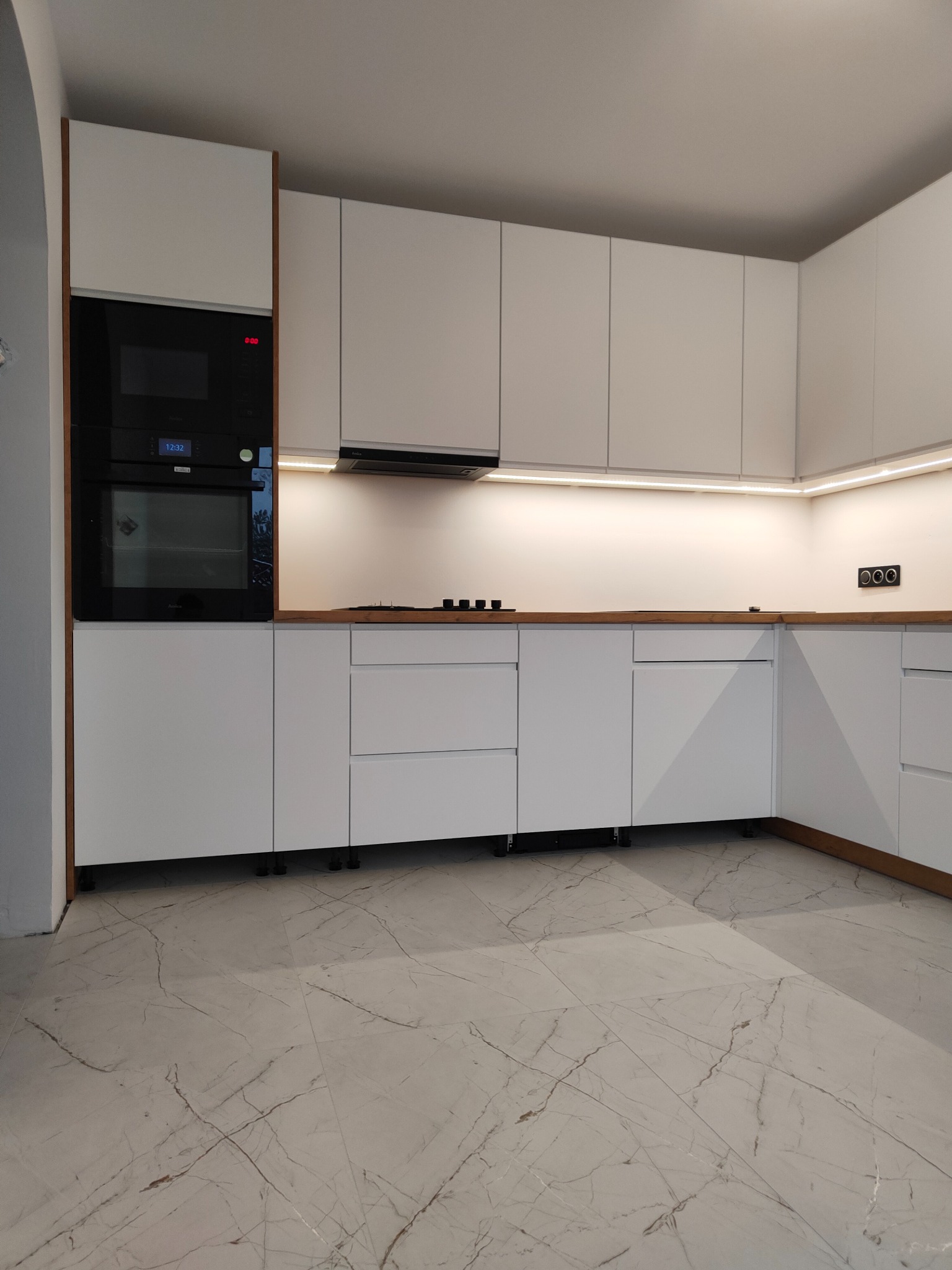 Nowoczesna, minimalistyczna zabudowa kuchenna z białymi frontami i drewnianym blatem, oświetlenie LED podszafkowe, widoczny piekarnik i płyta grzewcza, marmurowe płytki podłogowe.