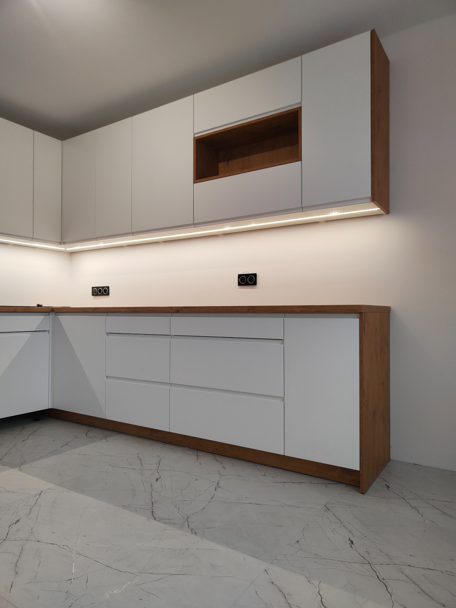 Nowoczesna, minimalistyczna kuchnia z białymi frontami i drewnianymi akcentami, oświetlona listwą LED podszafkową, na podłodze marmurkowy gres.