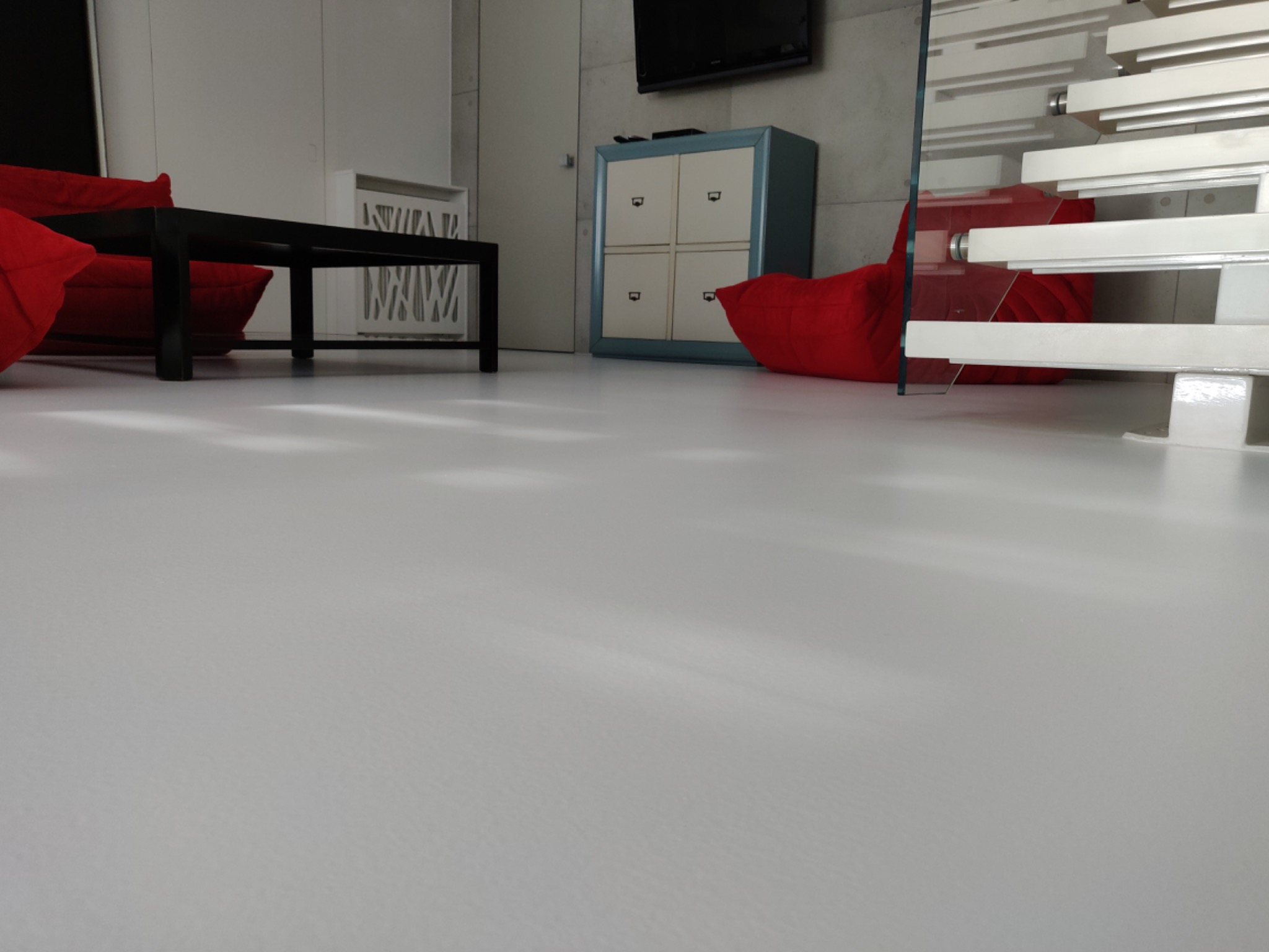 Biała posadzka żywiczna w minimalistycznym salonie z czerwonymi pufami, czarnym stolikiem kawowym i białymi schodami z elementami szklanymi.