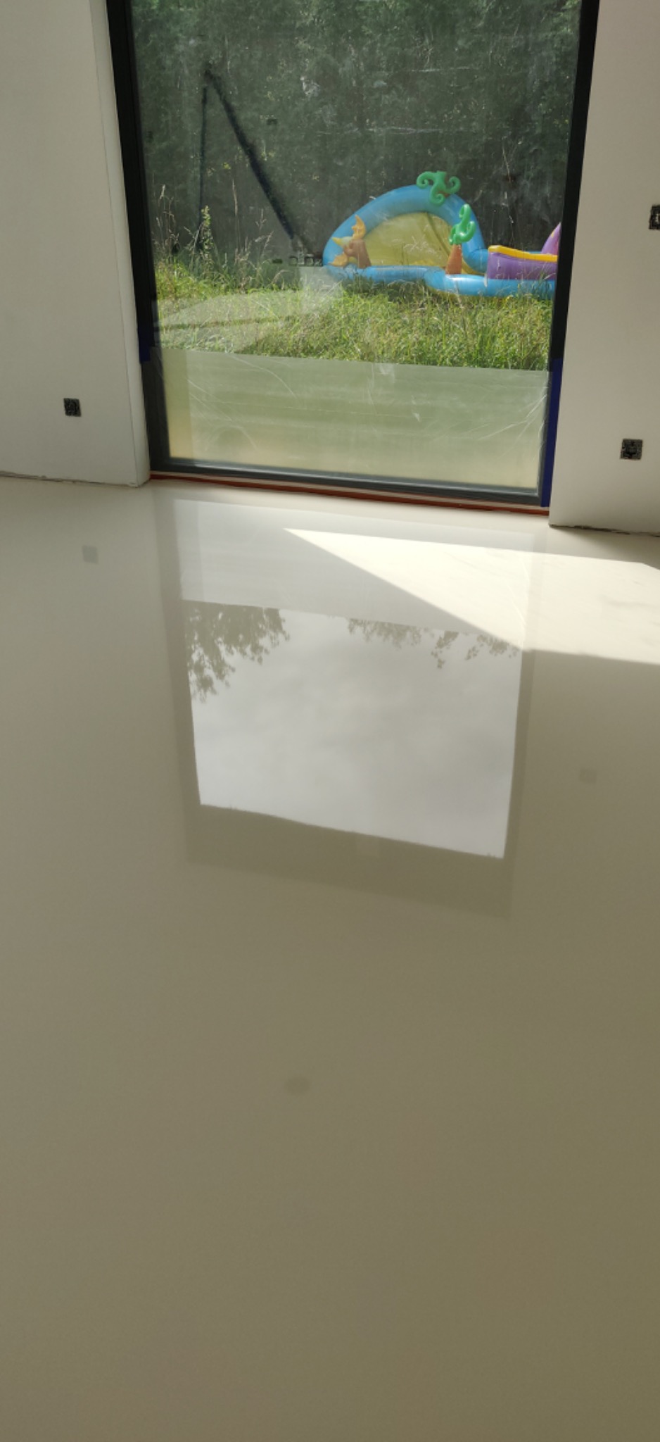 Błyszcząca, jasna posadzka z żywicy epoksydowej odbija widok z okna, gdzie widać trawnik i rozłożony basenik dla dzieci.