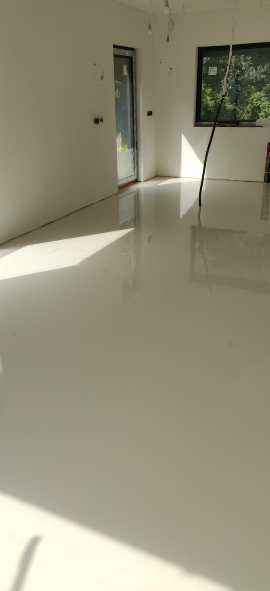 Biała, błyszcząca podłoga w nowym pomieszczeniu z dużymi oknami i niedokończonymi instalacjami elektrycznymi.