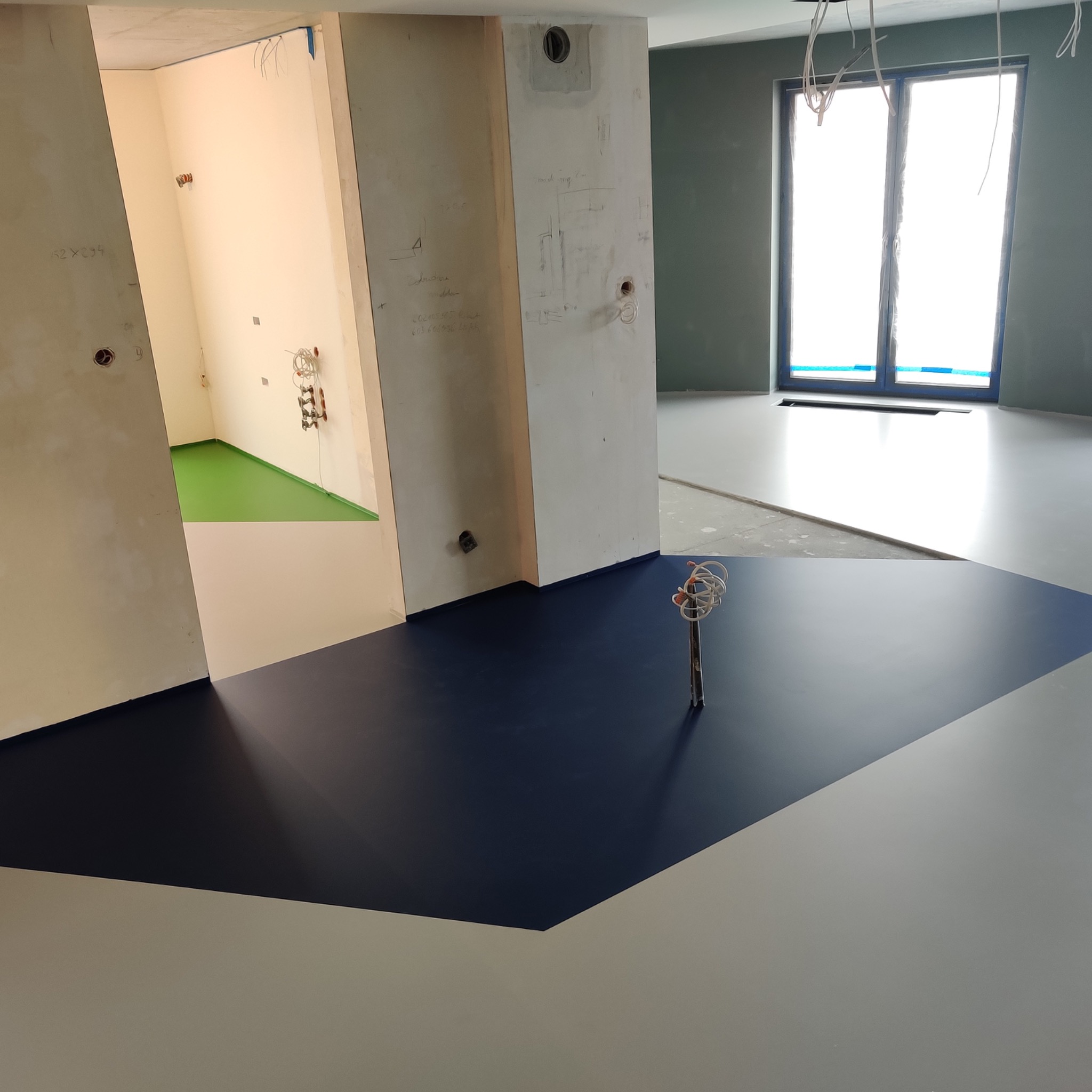 Wnętrze w trakcie wykańczania z dwukolorową, gładką posadzką w odcieniach szarości i granatu, widoczne kable wystające z podłogi oraz fragment zielonej posadzki w przyległym pomieszczeniu.
