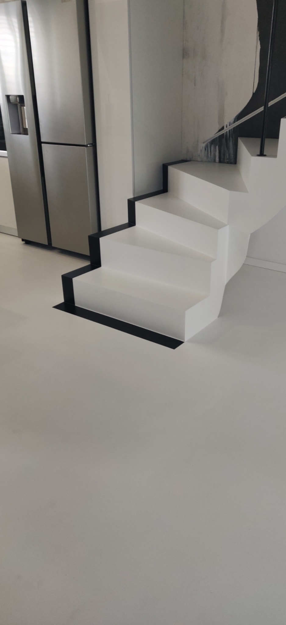 Białe schody z czarnymi obramowaniami na jednolitej, jasnej posadzce, obok srebrna lodówka w minimalistycznej kuchni.