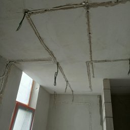 Mateusz Chrostowski - Surowy sufit i ściany w trakcie prac, widoczne białe rurki instalacji elektrycznej prowadzone w bruzdach, wystające przewody, oraz okno zabezpieczone folią.