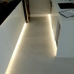 Nowoczesna kuchnia z białymi, błyszczącymi szafkami i podświetleniem LED pod szafkami, oświetlającym szarą podłogę z płytek.