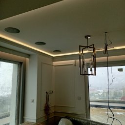 Mateusz Chrostowski - Nowoczesne wnętrze w trakcie wykańczania: podwieszany sufit z oświetleniem LED, głośniki sufitowe, designerska lampa wisząca, okno z widokiem na miasto, meble zabezpieczone folią.