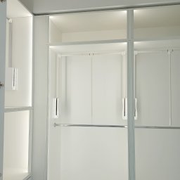 Mateusz Chrostowski - Wnętrze nowoczesnej, białej szafy wnękowej z oświetleniem LED, widoczne półki i drążki na ubrania.