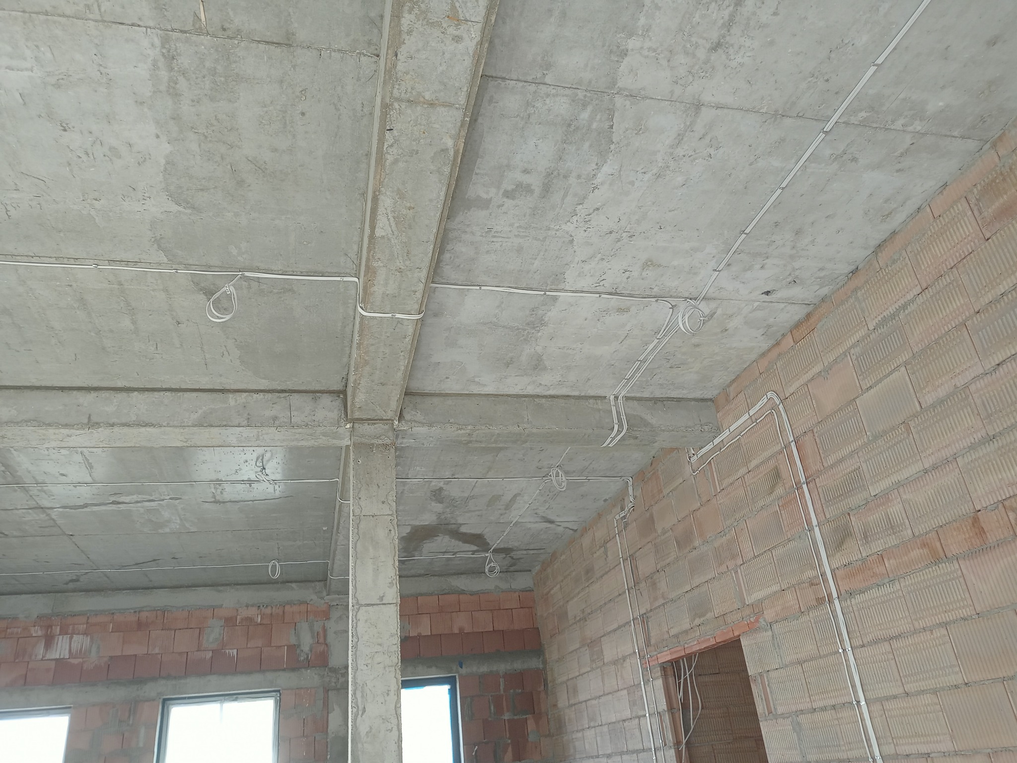 Surowy betonowy strop z widocznymi przewodami elektrycznymi w białych listwach, ceglana ściana w tle, stan surowy budynku.
