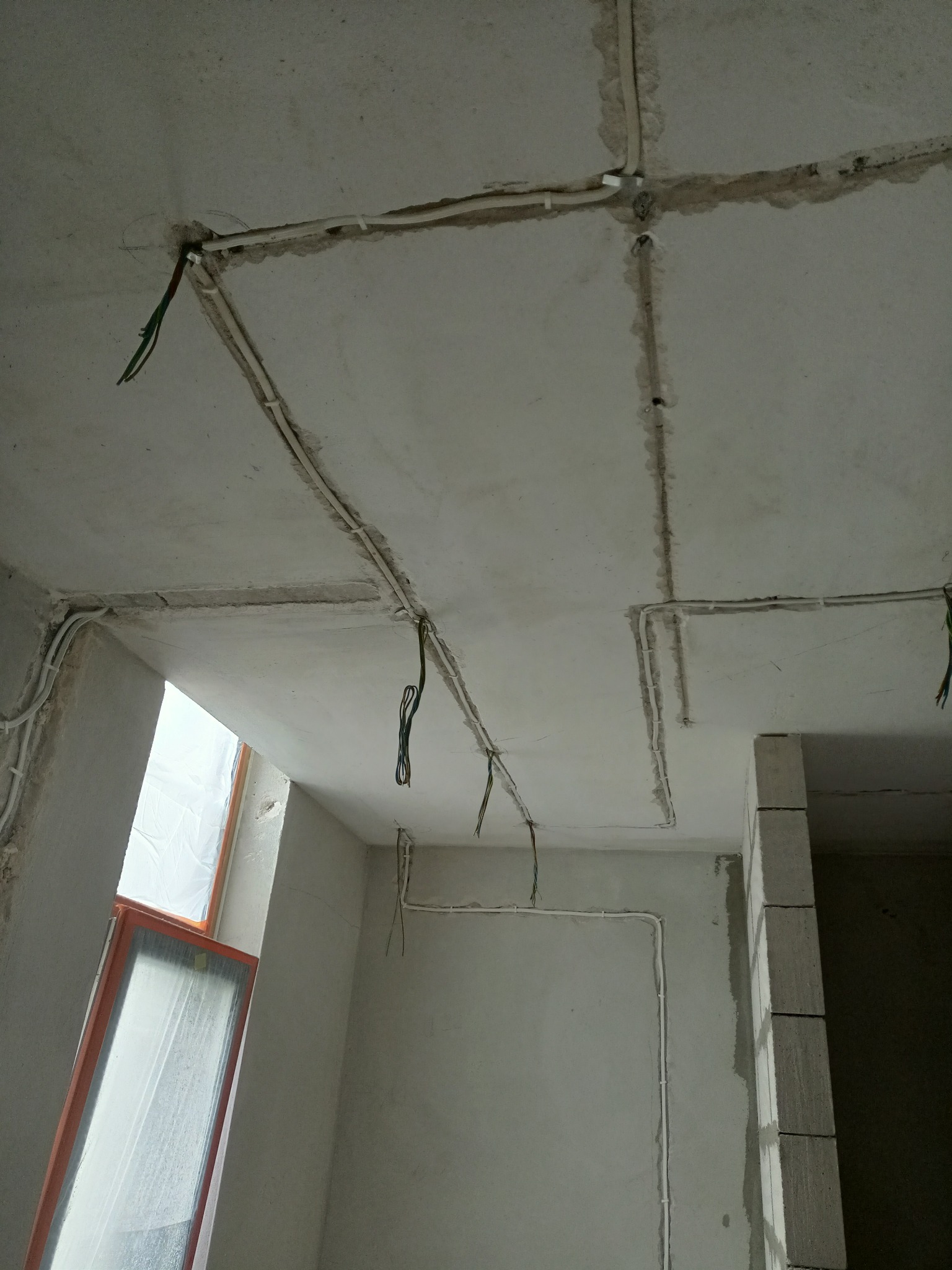 Surowy sufit i ściany w trakcie prac, widoczne białe rurki instalacji elektrycznej prowadzone w bruzdach, wystające przewody, oraz okno zabezpieczone folią.