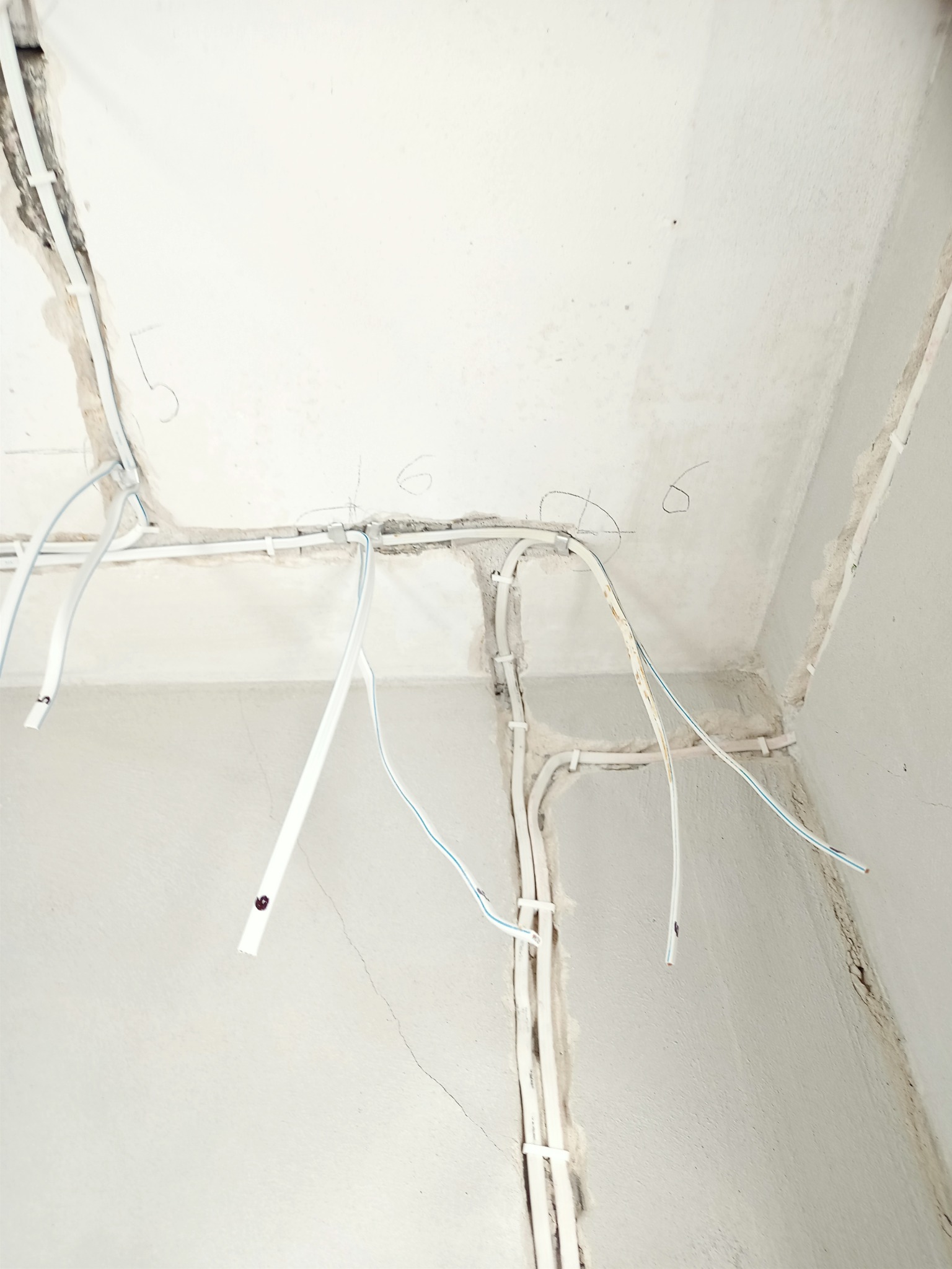 Surowe ściany i sufit w trakcie prac, z widocznymi przewodami elektrycznymi zamocowanymi do powierzchni i wiszącymi luźno, oraz numerami ołówkiem na ścianie.