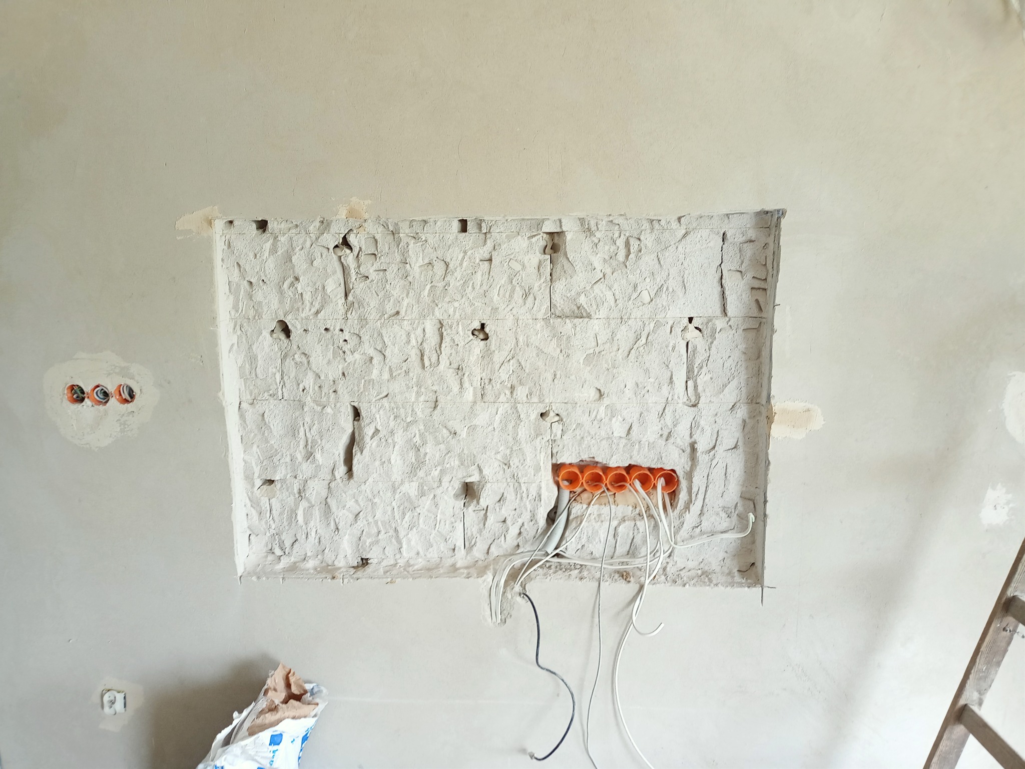 Ściana w trakcie prac przygotowawczych pod montaż instalacji elektrycznej: widoczne pomarańczowe rury instalacyjne z wystającymi przewodami, surowy tynk w miejscu planowanego gniazdka i drabina...
