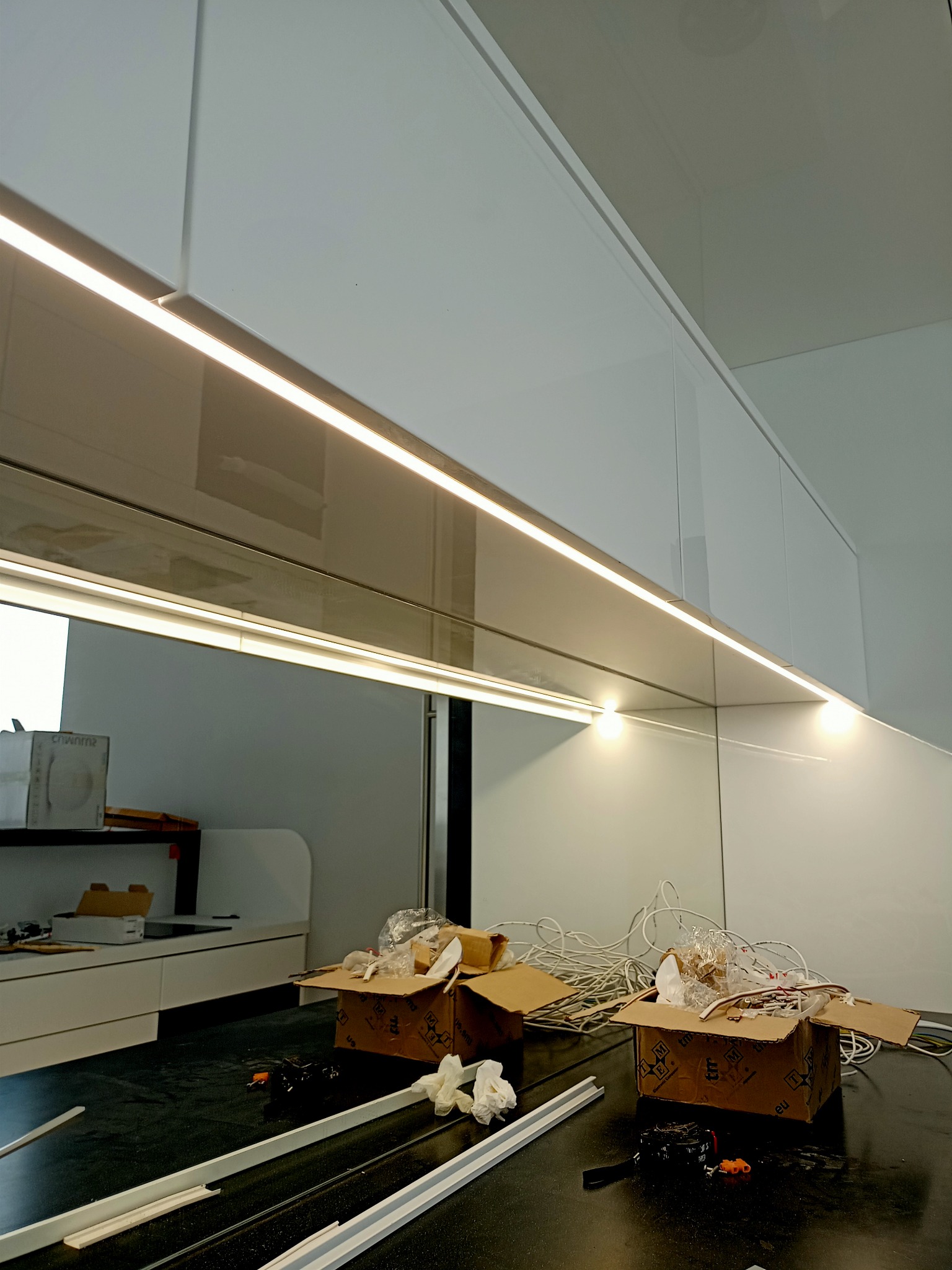 Białe szafki kuchenne z zamontowanym oświetleniem LED, widoczne kartony, kable i narzędzia sugerujące etap instalacji.