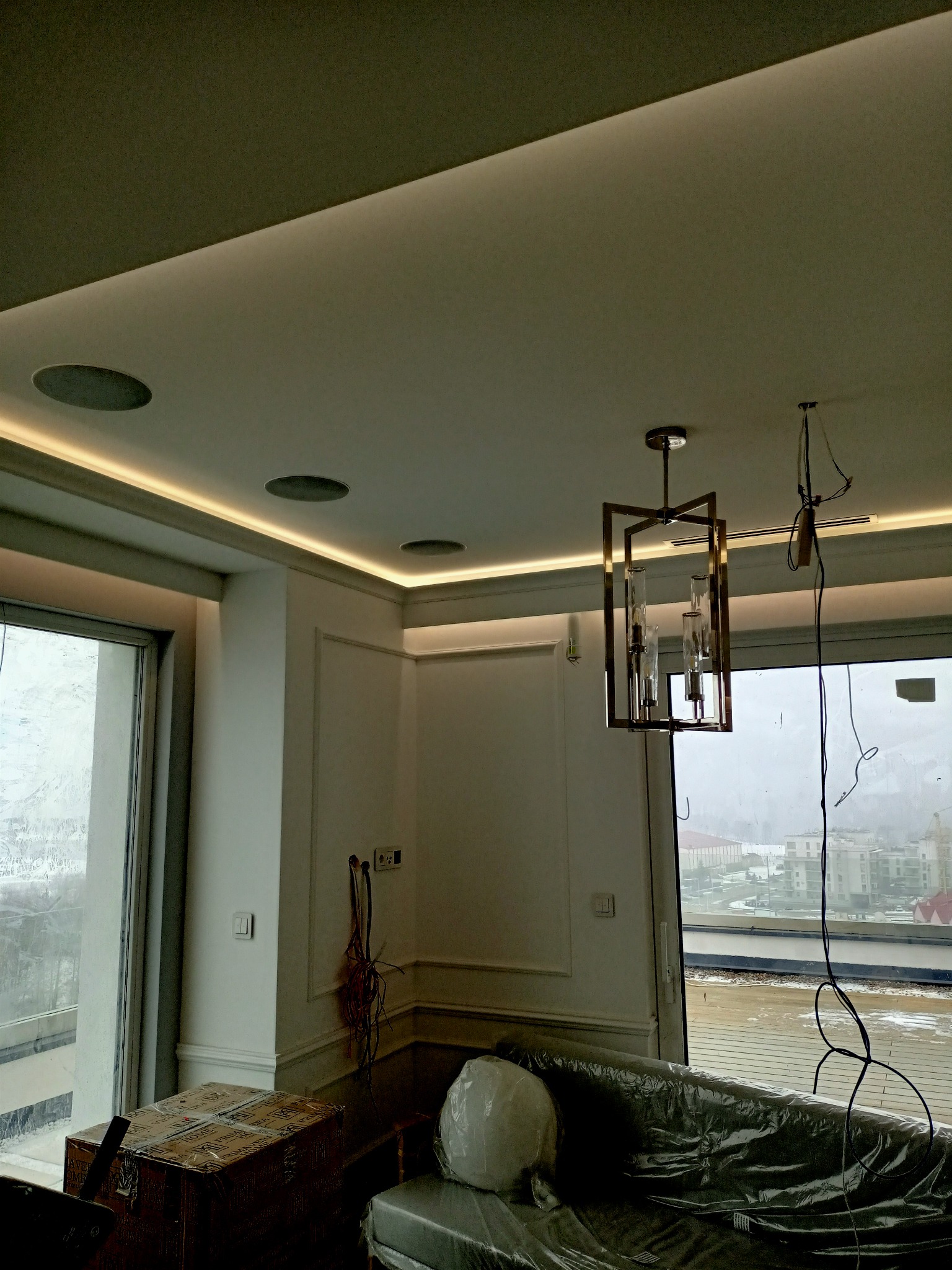 Nowoczesne wnętrze w trakcie wykańczania: podwieszany sufit z oświetleniem LED, głośniki sufitowe, designerska lampa wisząca, okno z widokiem na miasto, meble zabezpieczone folią.