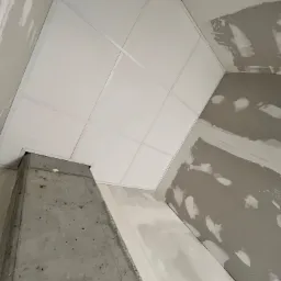 Sufit podwieszany z płyt gipsowo-kartonowych w trakcie montażu, widoczny surowy betonowy filar i ściana z widocznymi śladami szpachlowania.