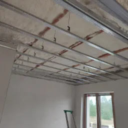 Poddasze w trakcie ocieplania pianką, widoczne metalowe profile sufitowe i drabina oparta o ścianę przy oknie.