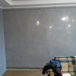 Świeżo pomalowana szara ściana z efektem dekoracyjnym, widoczne listwy przypodłogowe i sufitowe, na pierwszym planie fragment sofy z ubraniami.