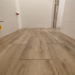 Ułożone panele podłogowe imitujące drewno w łazience, widoczne przyłącza wodne i fragment ściany z płytkami.