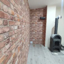 Ściana wykończona tapetą imitującą cegłę, z widocznym kominem i piecem wolnostojącym, na podłodze panele imitujące drewno.