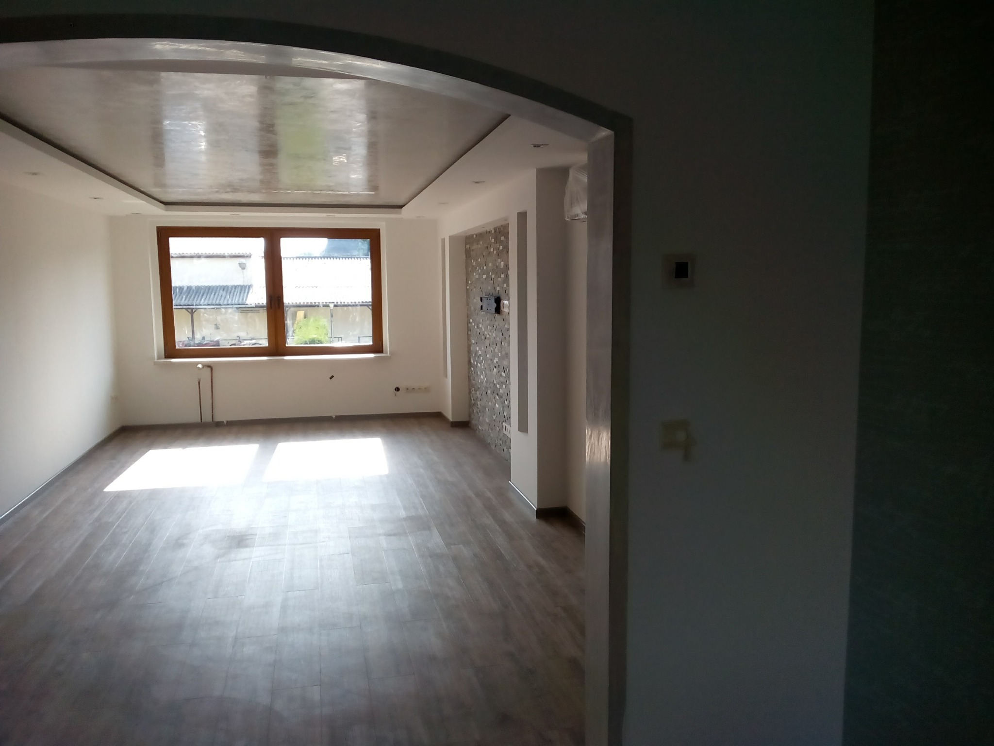 Jasne, puste wnętrze z panelami podłogowymi, błyszczącym sufitem podwieszanym i oknem z widokiem na budynki gospodarcze, ściana akcentowa z mozaiką.