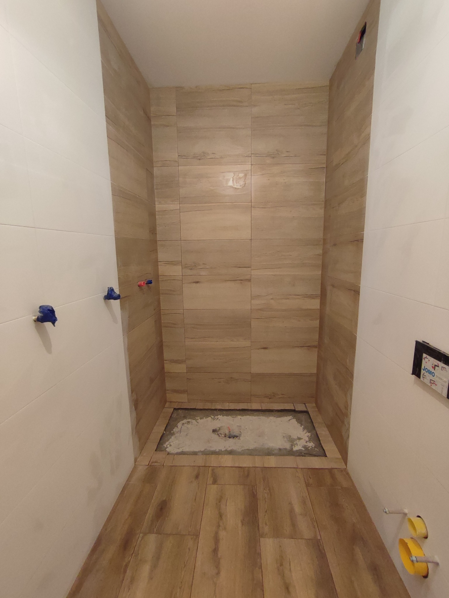 Wnętrze łazienki w trakcie prac: ułożone płytki imitujące drewno na podłodze i ścianach kabiny prysznicowej, widoczne wystające rury z niebieskimi i czerwonymi zaślepkami, niezakończone prace wokół...