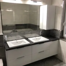 Nowoczesna łazienka z dwoma białymi umywalkami nablatowymi na szafce z szufladami, ciemny blat, lustro z oświetleniem i wisząca szafka.