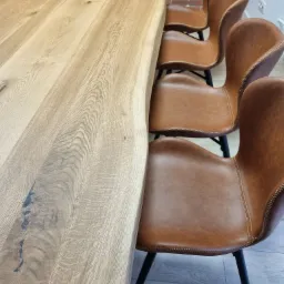 Blond drewniany blat stołu z naturalną krawędzią i widocznymi słojami, zestawiony z brązowymi skórzanymi krzesłami na czarnych metalowych nogach, na tle ceglanej ściany.