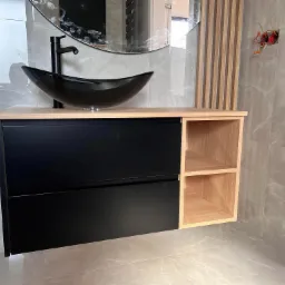 Nowoczesna łazienka z czarną, owalną umywalką na jasnym blacie, czarne szuflady i otwarte półki z jasnego drewna, szare płytki imitujące marmur, okrągłe lustro i drewniane lamele na ścianie...