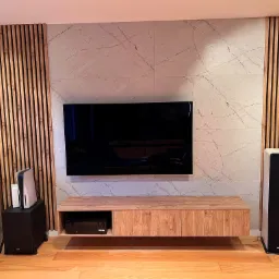 Nowoczesny salon z drewnianymi panelami lamelowymi po bokach marmurowej ściany, podwieszana szafka RTV z drewnianym blatem, telewizor na ścianie, głośniki podłogowe Teufel, konsola Playstation 5...