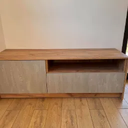 Niska szafka RTV z drewnopodobnym korpusem i szarymi frontami o fakturze betonu, umieszczona w rogu pomieszczenia na panelach podłogowych.